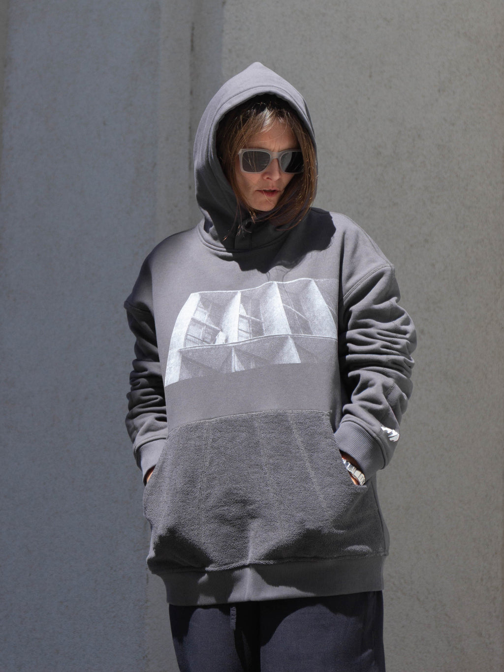 A person wearing a gray oversized hoodie with a large white geometric graphic on the chest, standing against a textured gray wall.