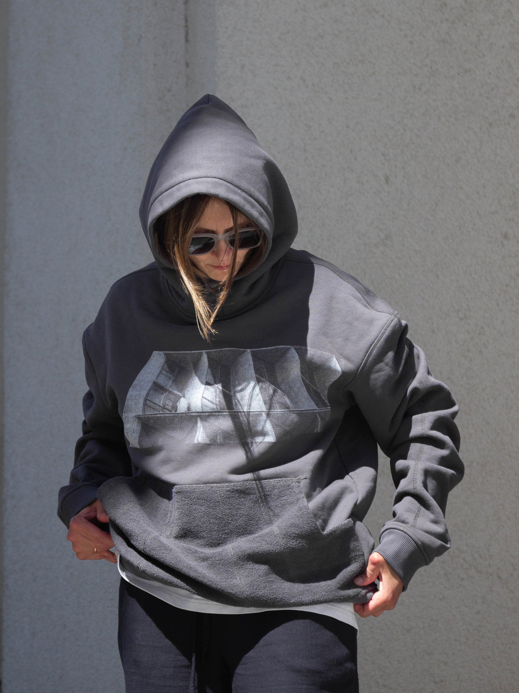 A person wearing an oversized dark gray hoodie with a brutalist print on the front, styled with dark pants and sunglasses, standing against a textured gray wall.